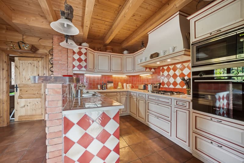 Open kitchen with wooden beams, white and red checkered backsplash, tiled floor, island, large range hood, oven, microwave, and various kitchen appliances