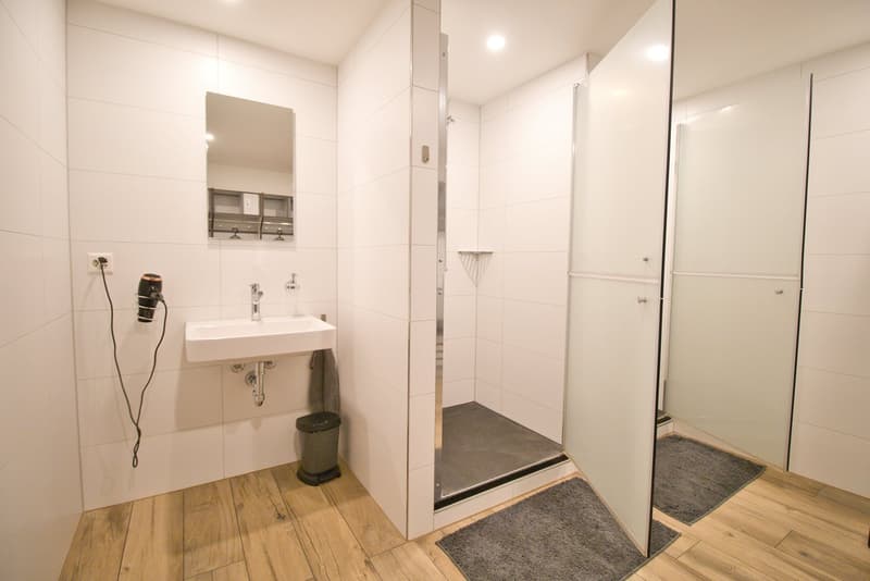 white tiles, wooden floor, white sink, mirror, shower, trash can, power outlet