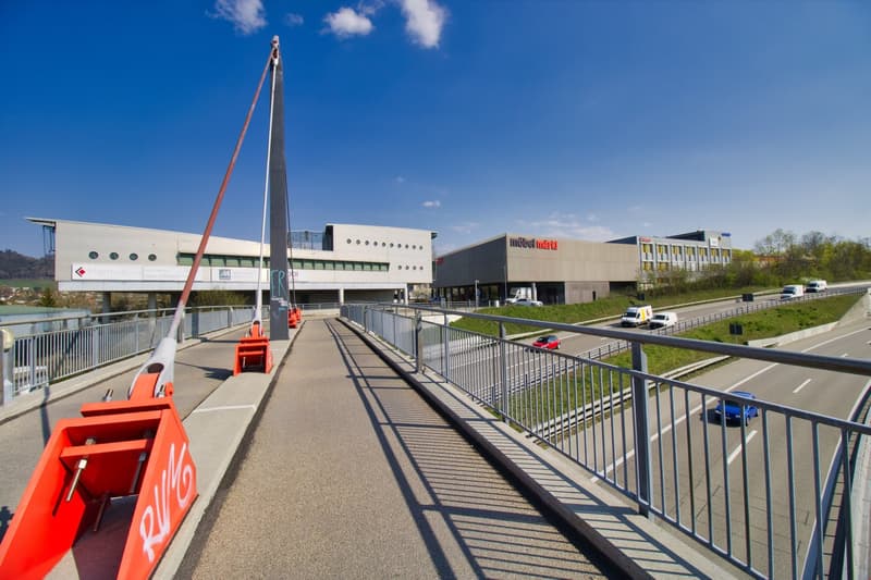 A large building with a modern design, featuring a large sign reading 'möbel mart', a bridge with metal railings, and a road with cars passing by.
