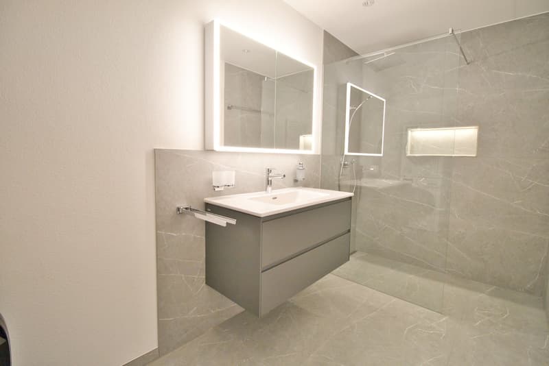 modern bathroom with white sink, gray vanity, glass shower enclosure, illuminated mirror, and built-in lighting