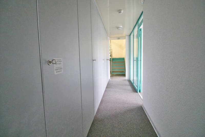 Empty hallway with carpeted floor, white walls, and turquoise doors. Stairs leading up to the next floor.