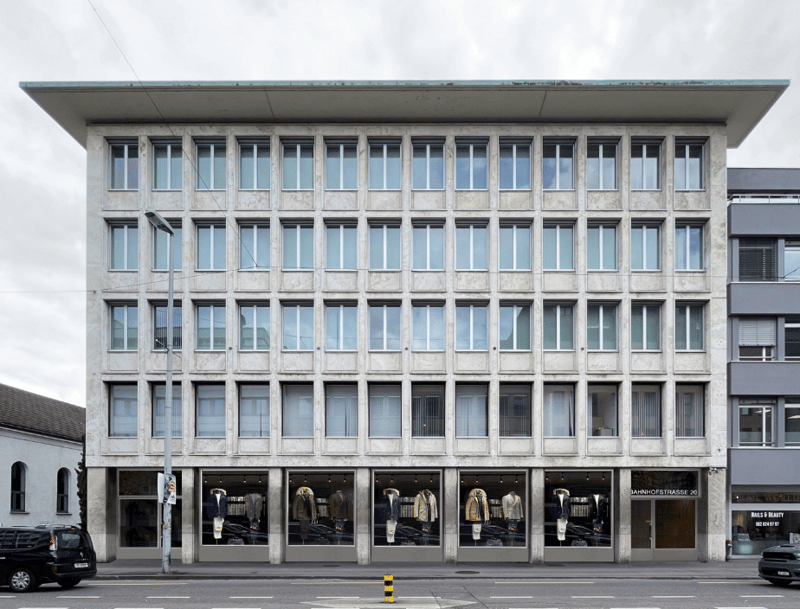 Modern architectural design with multiple floors, concrete exterior, large windows, multiple storefronts on the ground floor, street-level parking visible, multiple parked cars in front.