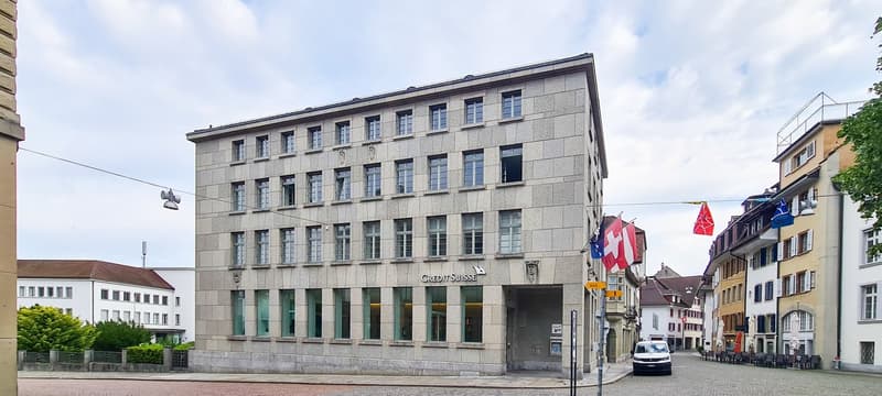 A commercial building with glass windows, a brick front, and a large sign reading 'Credit Suisse'. A white van is parked in front.