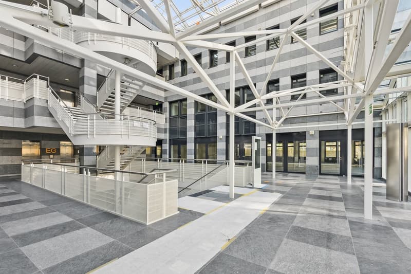 The image shows a large, open-plan interior space with a high, glass ceiling and a modern, industrial-style design. The space features a complex network of metal beams and supports, creating a visually striking architectural structure. The floor is covere