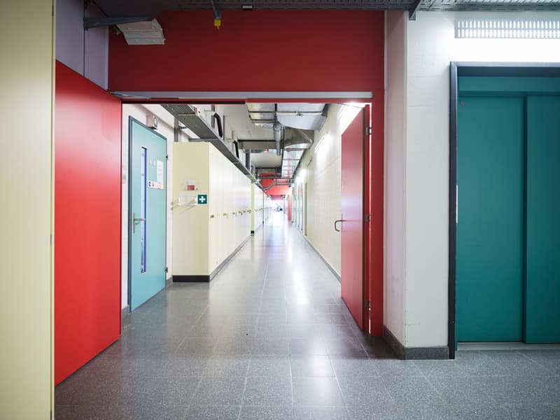 Long and narrow corridor, red and green doors, shiny tiles, closed doors on both sides.