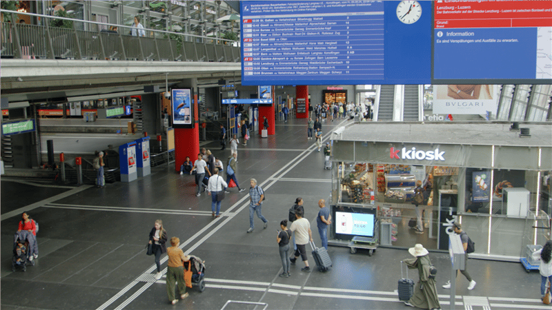 Airport terminal with passengers walking, kiosks, digital information board, escalators, and luggage carts.