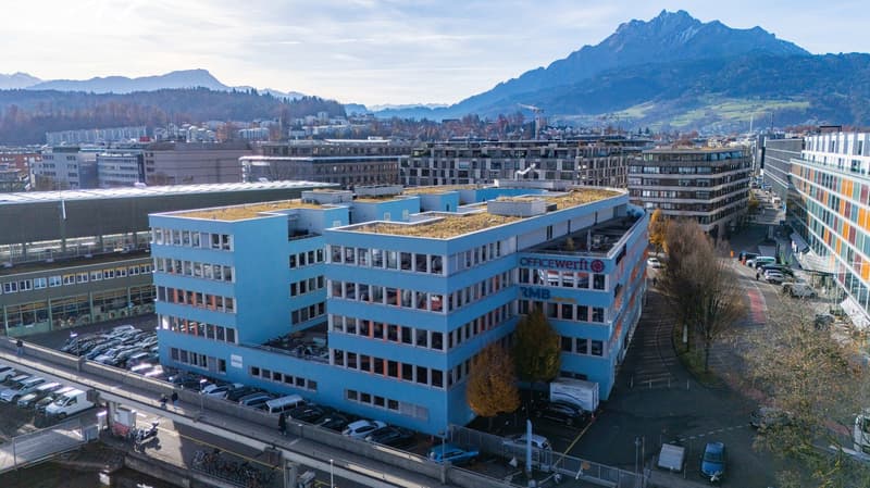 Blue building with green roof, multiple stories, office space, exterior view, several cars parked in front, city view in the background.