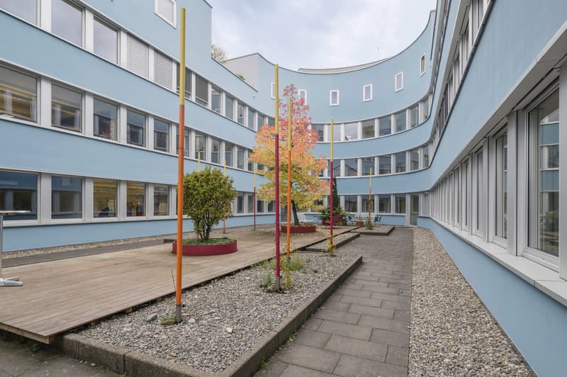 Multi-storey building with multiple windows, paved walkway, shrubs, and poles in the center
