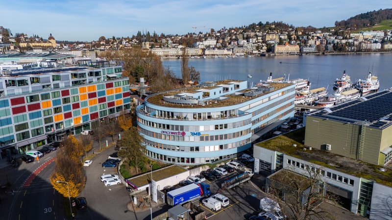 An aerial view of an office building, featuring several floors with multiple windows, surrounded by trees and a busy street with multiple cars parked in front. Other buildings, including another office building, are visible in the distance, along with a l