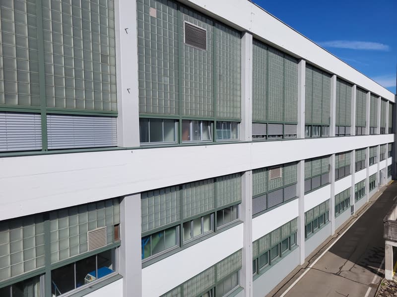 A commercial building with a glass wall facade, white exterior, and multiple floors. Several windows and ventilation grilles are visible.