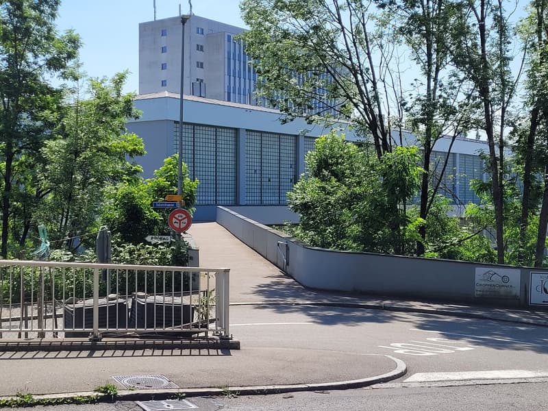 A two story building with glass panels in the front. On the right are multiple trees. On the left, there is a metal railing with garbage cans placed on the side. In the back is a walkway with fences on the sides.