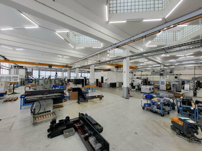 Large industrial workshop space with high ceilings, overhead cranes, and various machinery and equipment such as metal fabrication tools, workbenches, and storage racks. The space appears to be used for manufacturing or industrial production.