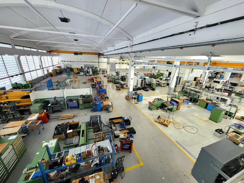 Large industrial workshop with various machinery, tools, and equipment. The space has high ceilings, overhead cranes, and is filled with workbenches, storage shelves, and other industrial items.