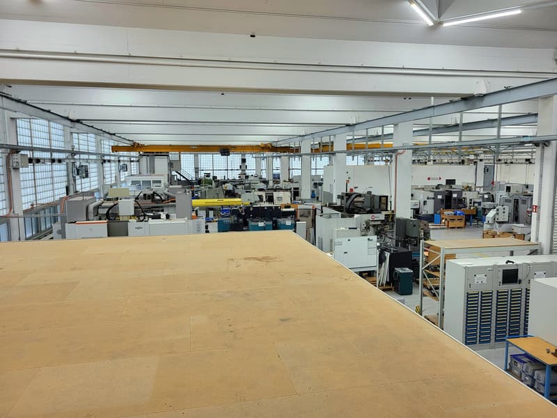 Large industrial workshop space with various machinery, equipment, and storage racks. The space has high ceilings, concrete floors, and overhead cranes or hoists. The workshop appears to be used for manufacturing or assembly of various products.