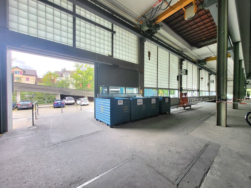 Large open warehouse space with concrete floor, high ceiling, and glass windows overlooking a parking area outside. There are several blue tool chests or storage units visible in the space.