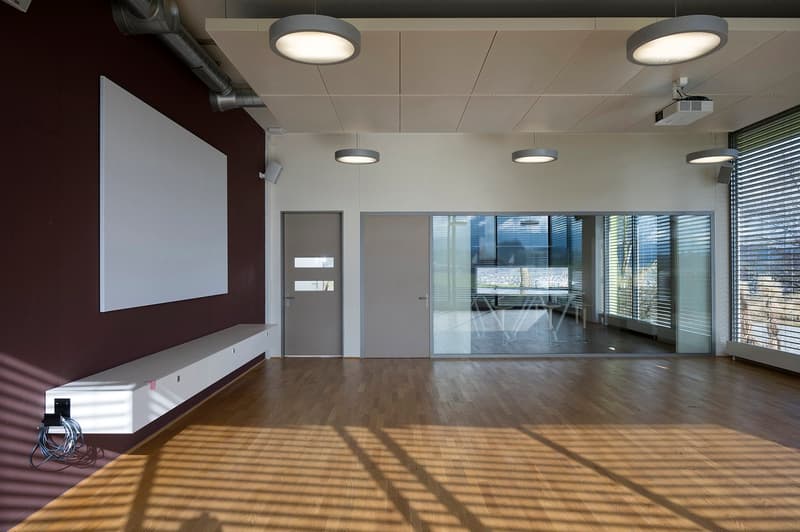 Large open space with hardwood flooring, white walls, and recessed lighting. There is a projector screen mounted on the wall and glass doors leading to an outdoor area.