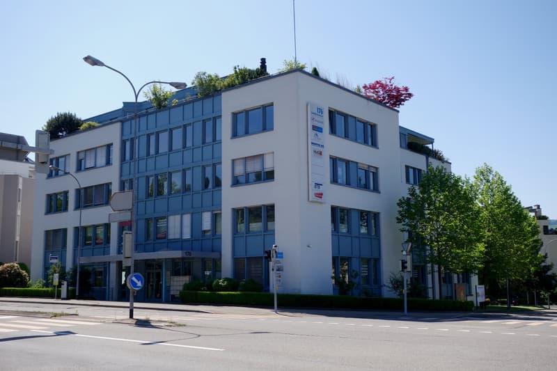 A multi-story office building with a modern, glass-fronted design. The building has several floors and is surrounded by trees and greenery. There is a road in front of the building with a traffic sign visible.