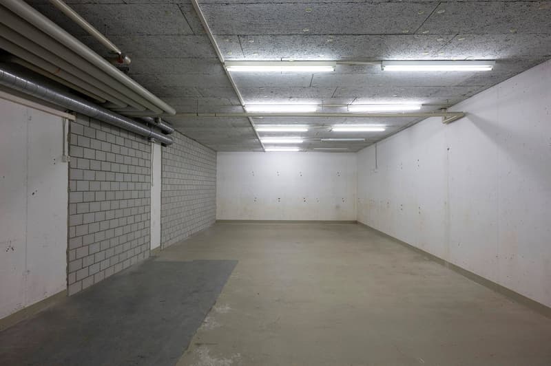 Empty underground parking area with white walls, brick pillar, and multiple fluorescent lights.