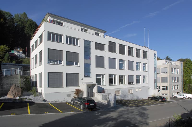 A multi-story office building with a modern, minimalist design. The building has large windows, a flat roof, and a parking lot in front. The exterior is primarily white with some gray accents. The building appears to be situated in a hilly, forested area