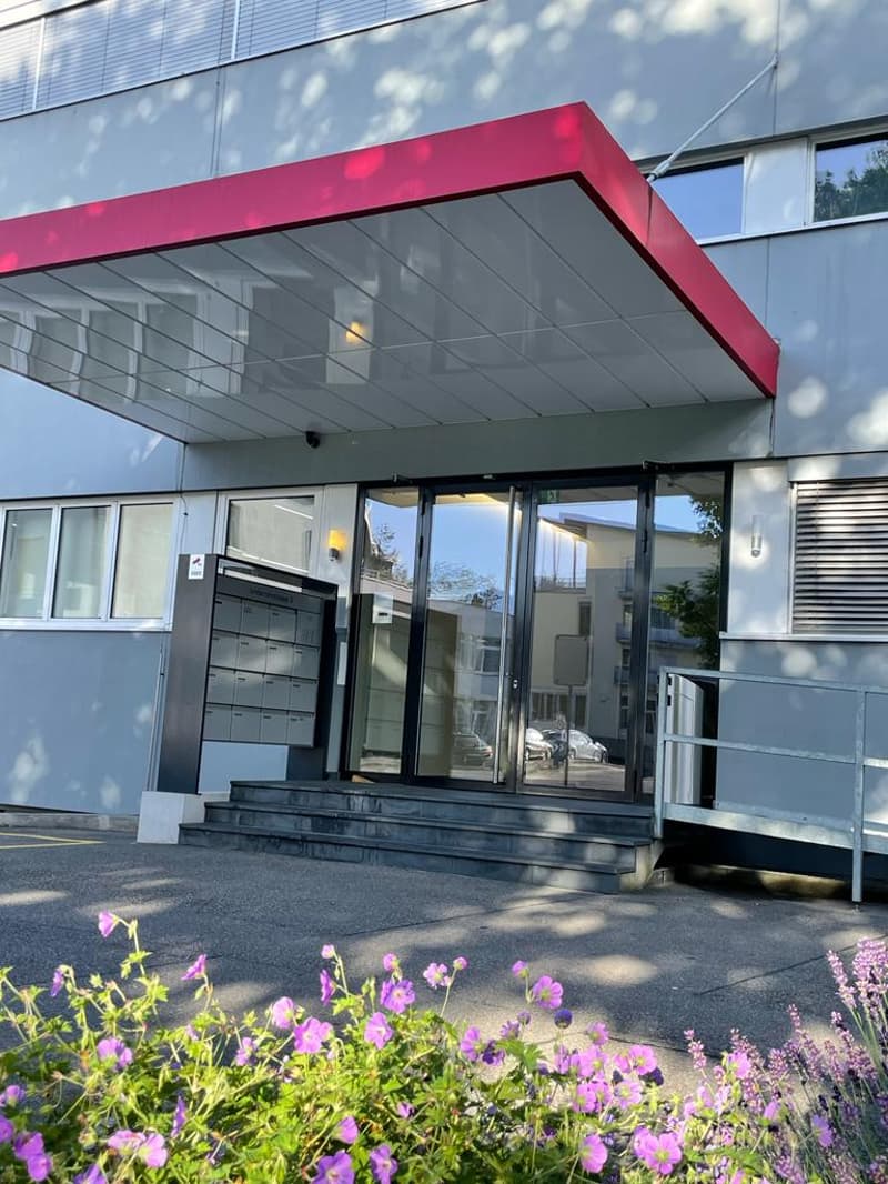 Modern commercial building with a red canopy over the entrance, glass windows, and a flower garden in the front