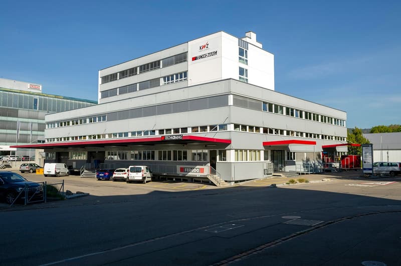 Multi-story commercial building with the Tecnomag logo, parking spaces in the front, and various vehicles parked outside the building.