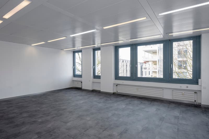 An empty office room with gray flooring, white walls, three windows with gray frames, and ceiling lights