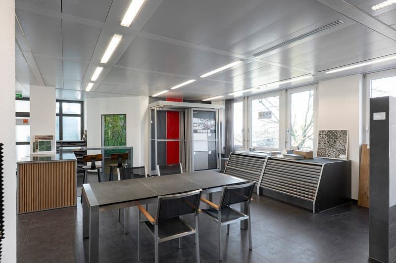 Modern room with gray tables, chairs, two doors, two windows with curtains, a wooden counter with display glass, shelves with artwork, ceiling with lights.