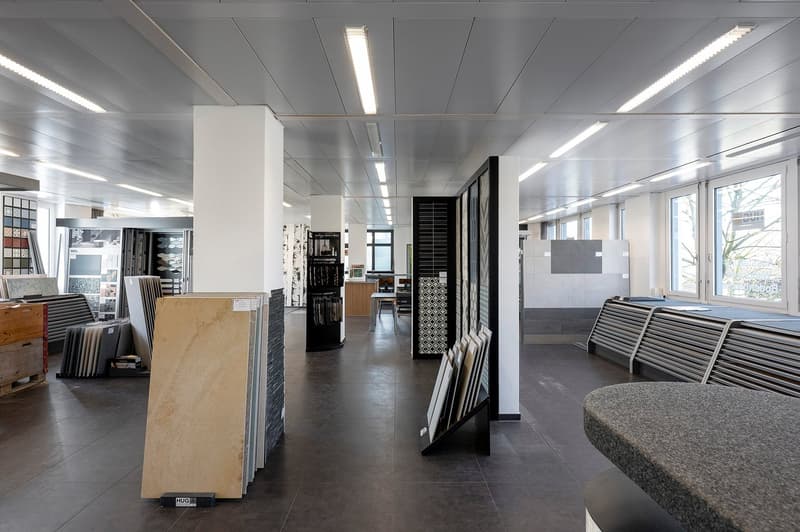spacious room, gray flooring, illuminated with bright overhead lights, displays of tiles and building materials, central aisle, display stands with sample tiles, modern design, industrial atmosphere