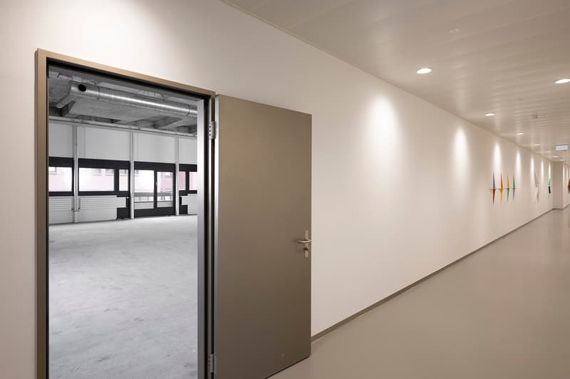 Long hallway with white walls, gray floor, exposed pipes, glass doors, windows, ceiling lights, colorful wall decorations