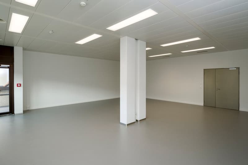 empty room, white walls, gray flooring, central column, recessed ceiling lights, door, glass panel on left, emergency light
