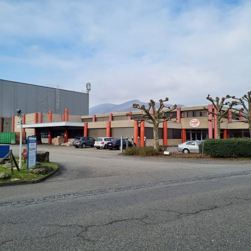 Industrial building with glass windows, parked cars in front, red and white colored, green container behind