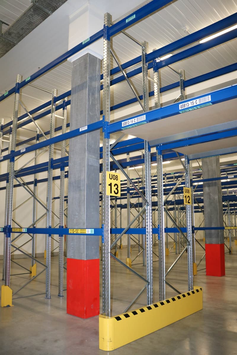 Warehouse with metal shelving units, yellow and red pillars, yellow and black bumpers, white ceiling, and barcodes on the shelves.