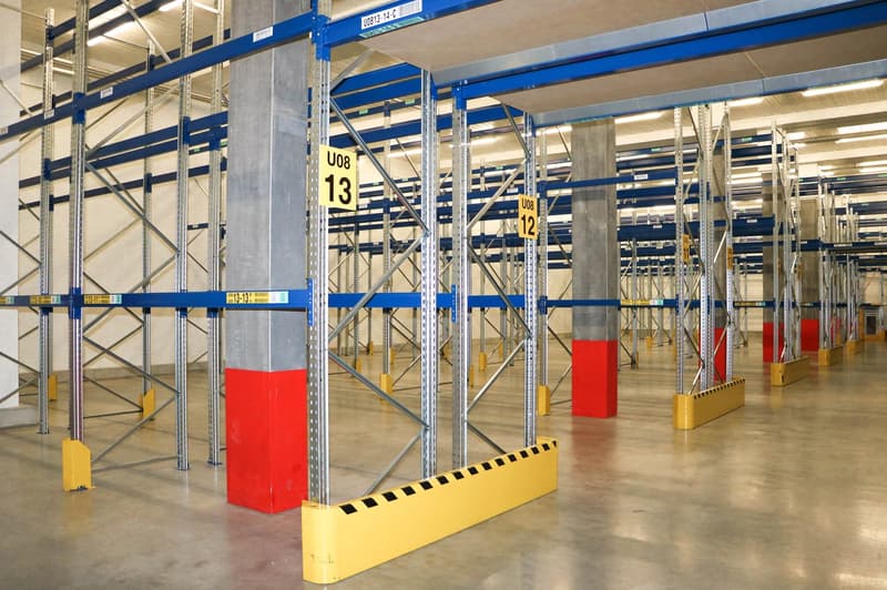 Warehouse with metal racks, yellow and red bumpers, yellow and red pillars, yellow and red signs, concrete floor, metal beams, and lights on the ceiling.