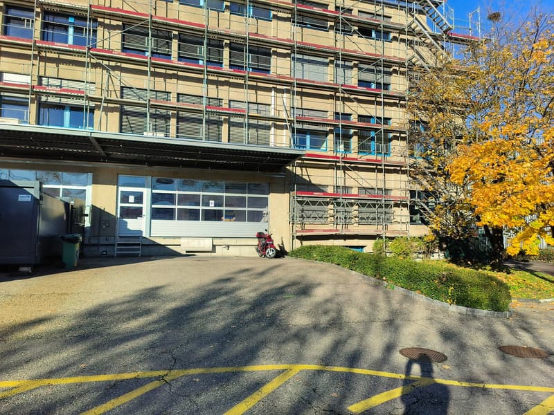 Building with red brick, multiple floors, several windows, under renovation, with some scaffolding, empty parking area in front, and a scooter
