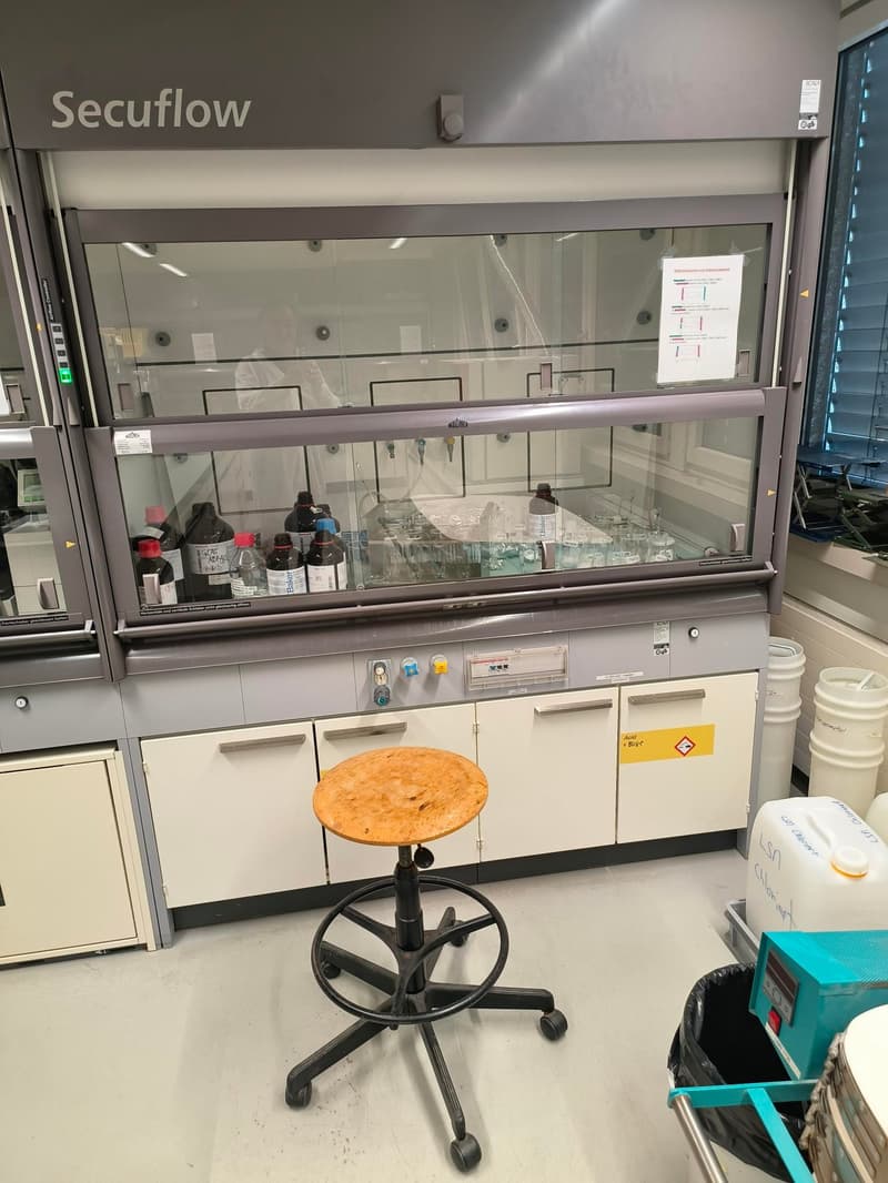 Secuflow work bench, glass cabinet with chemicals, round wooden stool, storage containers, white drawers, and safety warning signs