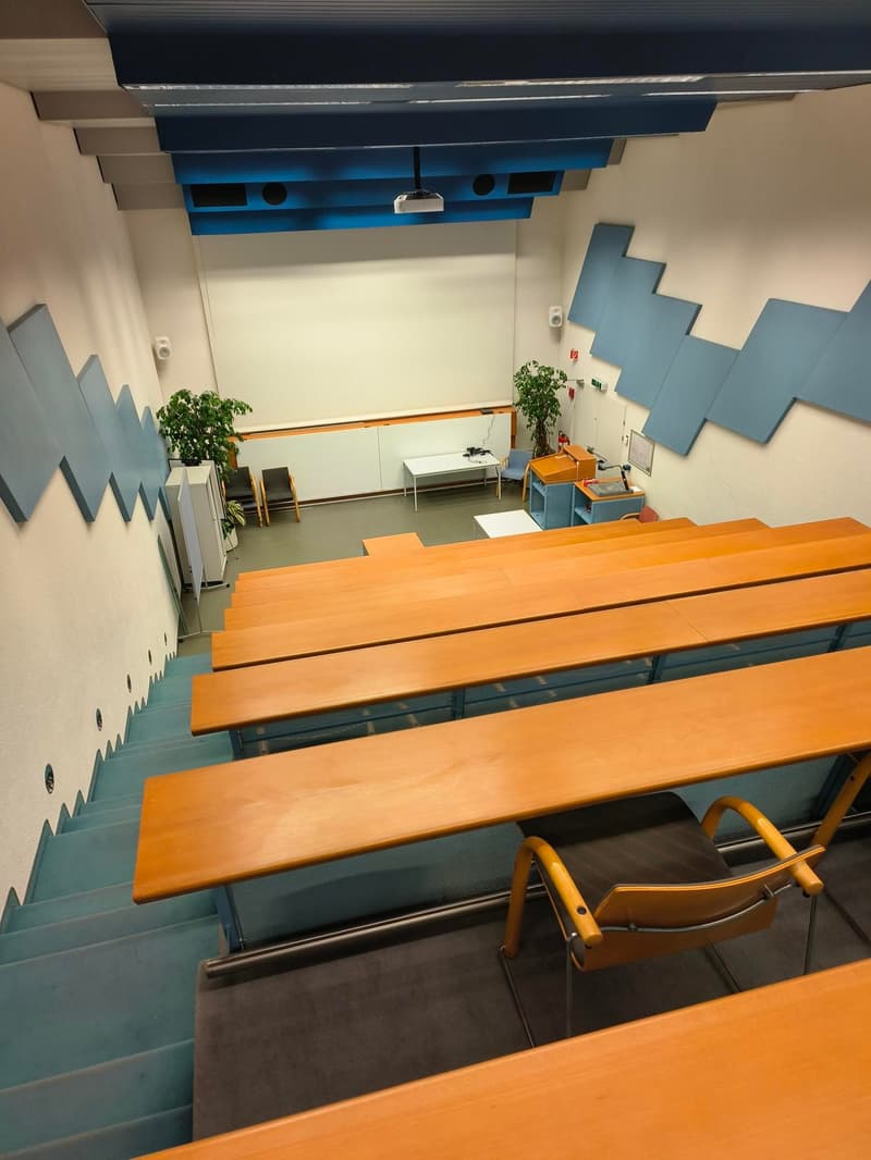 Wooden desks and chairs, white walls, blue patterned walls, staircase, metal railings, plants, desk, projector.