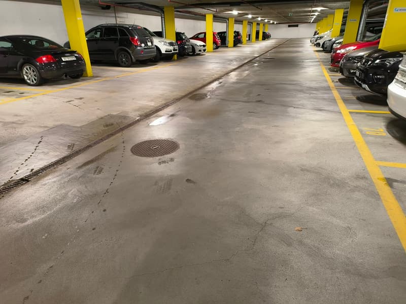 Underground parking lot with multiple cars parked, yellow and white markings, drain in the center, yellow pillars
