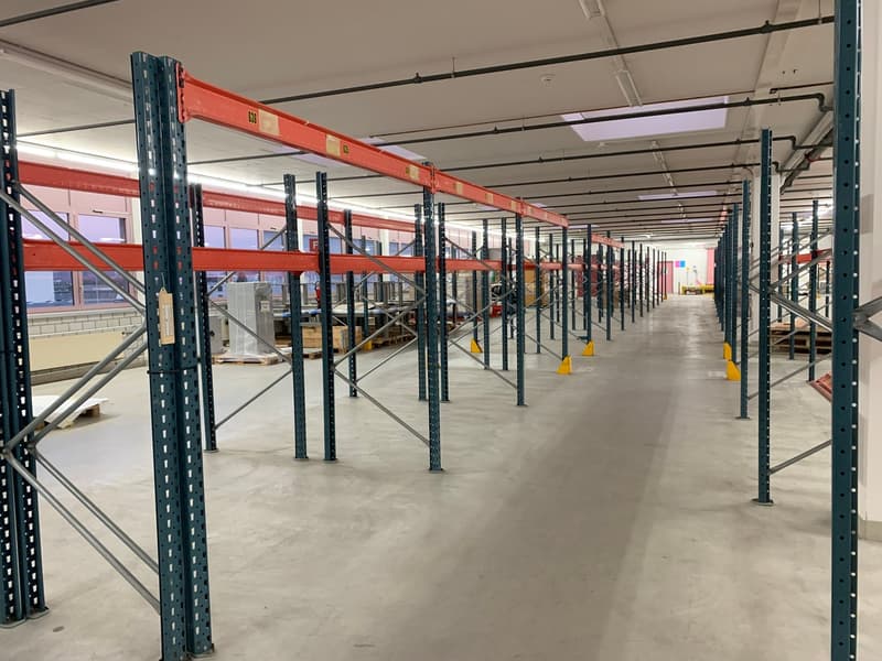 Warehouse with metal shelving units, yellow safety bollards, industrial lighting, empty, paved floor.