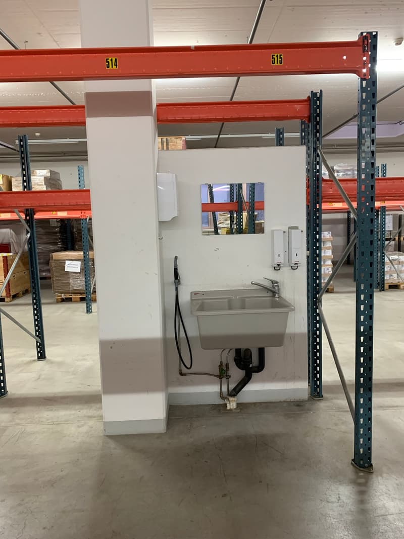 A room with concrete flooring, metal shelves, and a wash sink, mirror, and dispensers. There is an open area to the left.