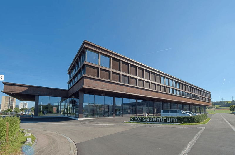 modern office building, glass facade, spacious parking area, brown facade, cars parked in front, trees and bushes, sign with 'Reisezentrum'