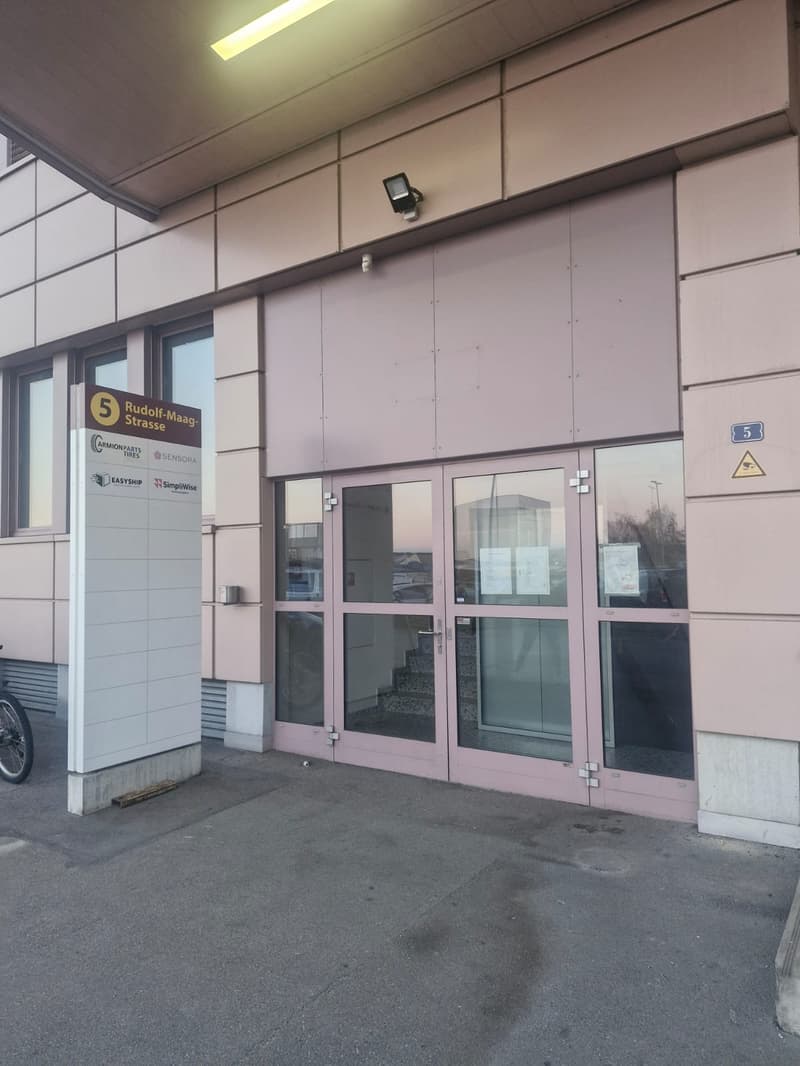An entrance to a building with glass doors, a sign indicating the Rudolf-Maag Strasse, and a surveillance camera mounted on the wall.