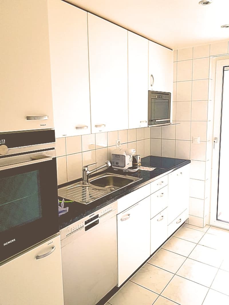 Modern kitchen with white cabinets, black countertop, built-in oven and dishwasher, tiled backsplash, and tile flooring.