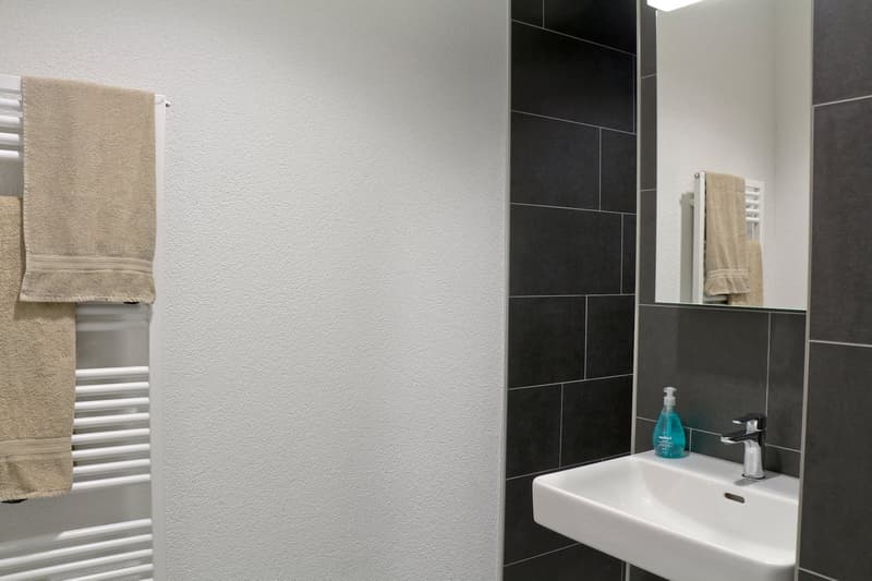 modern white sink, faucet, soap, mirror, towels on radiator, black tiled wall