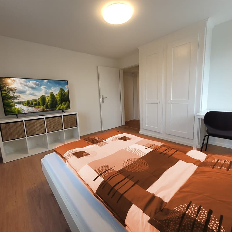 The bedroom is furnished with a bed, a TV on a shelf, a desk and chair, and white wardrobes.