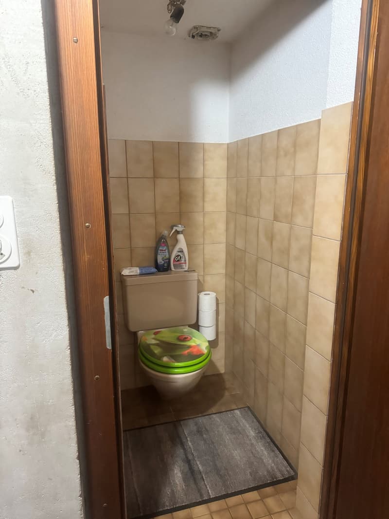 A small, tiled bathroom with toilet, tile flooring, and a single wall with a power outlet.