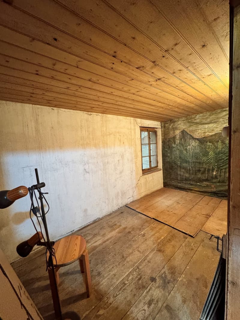 Room with wooden floors, white walls, wooden ceiling, wooden chair, tripod with lights, and a mural on the wall next to a window.