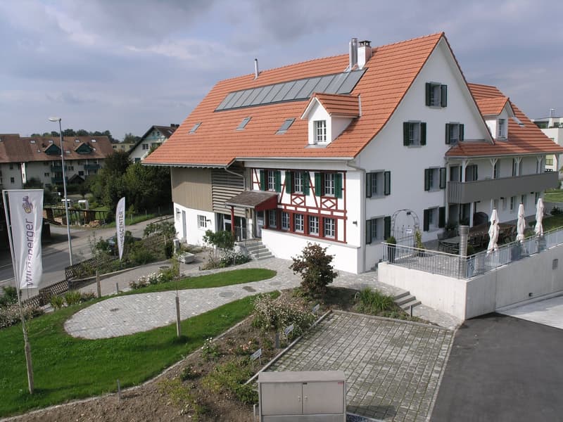 2 story house, white exterior, red roof, solar panels, green trim, large windows, raised ground floor, garden, pathway, outdoor seating