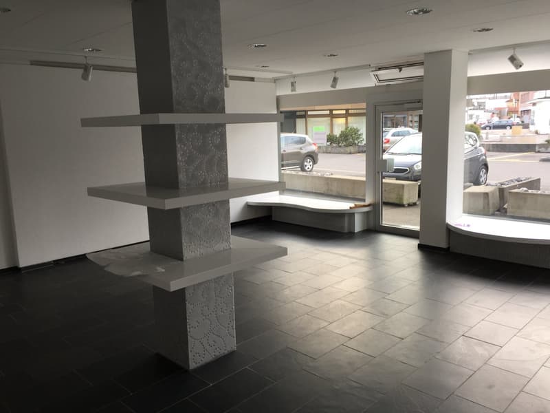 Empty room with white walls, black tiles, central column with shelves, glass doors, concrete bench, mounted lights.