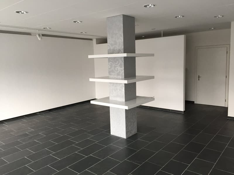 Empty room with white walls, black tiled floor, central decorative column, white shelves, ceiling lights