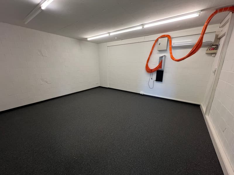 This image shows a small room with white brick walls, a carpeted floor, and overhead lighting. There is an orange cable running along the wall, but no other visible features.